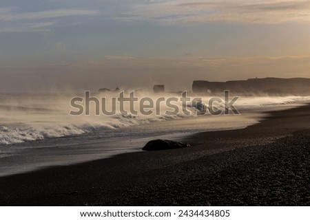 Sneaker waves crushing against Reynisfjara Black Sand Beach coast, with strong winds blowing water and volcanic black sand, sunset, extreme weather, Iceland. 