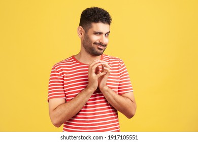 Sneaky cunful man with beard in striped red t-shirt drumming fingers planning devious pranks, thinking over revenge plan, scheming. Indoor studio shot isolated on yellow background