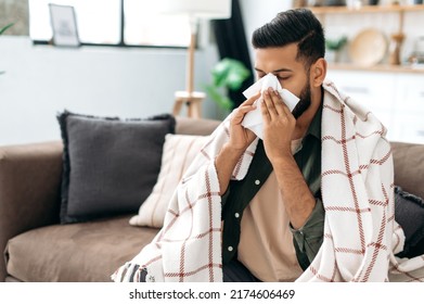 Sneezing, runny nose, cough. Ill arabian or indian young man, sits on the couch at home under a plaid, sneezing in a napkin, feels weak, unwell, needs treatment and medicines - Powered by Shutterstock