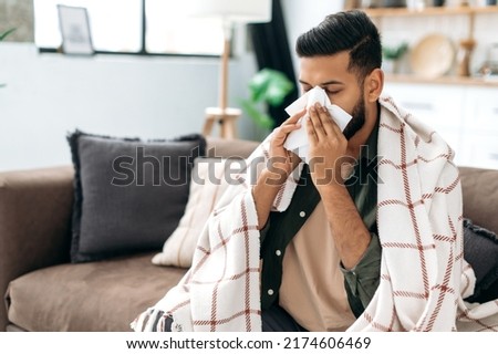 Sneezing, runny nose, cough. Ill arabian or indian young man, sits on the couch at home under a plaid, sneezing in a napkin, feels weak, unwell, needs treatment and medicines