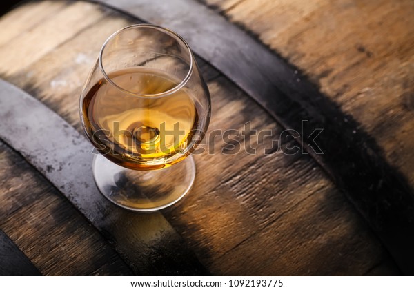 Snifter glass of brandy standing on an oak barrel\
in a cellar