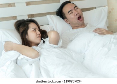 Snoring Asian Man And Young Asian Young Woman. Couple Sleeping In Bed. Young Girl Can't Sleep Because Of Her Man's Snoring. Man Snoring While His Wife Is Covering Ears With The Pillow.