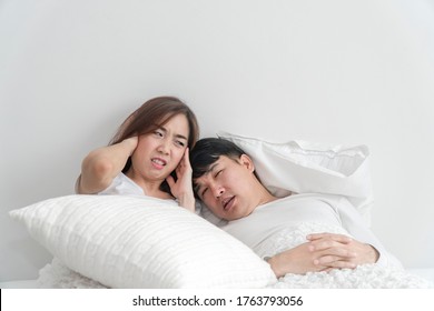 Snoring Asian Man And Young Asian Young Woman. Couple Sleeping In Bed. Young Girl Can't Sleep Because Of Her Man's Snoring. Man Snoring While His Wife Is Covering Ears With The Pillow.