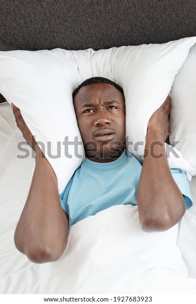 Snoring, insomnia, bad sleep, loud neighbors. Upset\
tired young african american man freaking out, covers his ears with\
pillow so as not to hear noise in bedroom, vertical, top view, copy\
space
