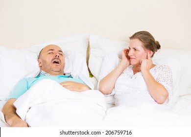 Snoring Man. Couple In Bed, Man Snoring And Woman Can Not Sleep, Covering Ears With Hands For Snore Noise. Middle Aged Couple, Attractive Woman, Caucasian Man Sleeping In Bed At Home. Face Expressions