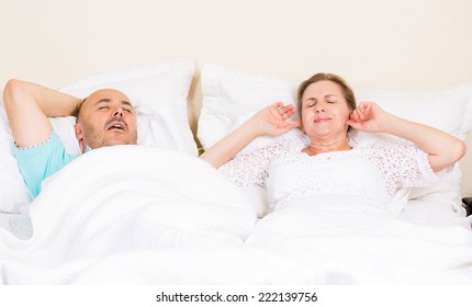 Snoring Man. Couple In Bed, Man Snoring And Woman Can Not Sleep, Covering Ears With Hands For Snore Noise. Middle Aged Couple, Attractive Woman, Caucasian Man Sleeping In Bed At Home. Face Expressions