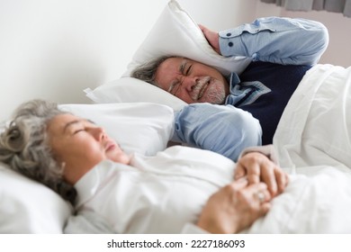 Snoring woman. Asian elderly couple sleeping on bed. Elderly Asian man having trouble sleeping squeezes white pillow around ears for peace and quiet - Powered by Shutterstock