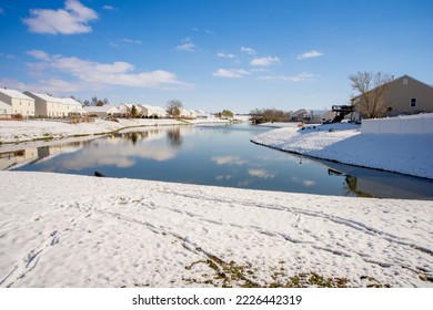 Snow around a lake in winter. Houses line the shore. Footprints can be seen in the snow. The sky is bright blue with puffy clouds. The water is a dark blue green. - Shutterstock ID 2226442319
