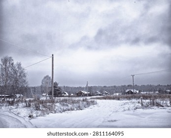 Snow blankets the path and fields in a rural area with houses visible in the distance under a gray sky during winter.