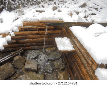 Snow blankets a rustic log spring shelter in the Czech mountains, where a metal spout pours water onto wet stones. Overcast winter light, conifer branches, and ice crusts shape a quiet scene.