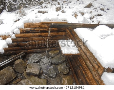 Snow blankets a rustic log spring shelter in the Czech mountains, where a metal spout pours water onto wet stones. Overcast winter light, conifer branches, and ice crusts shape a quiet scene.