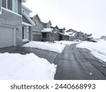 Snow blankets a suburban street, where houses stand in quiet repose and driveways reveal the mornings labor against the winter relentless drifts.