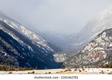 Snow blowing between mountains as springtime comes to Colorado.  Trees in the foreground of the Rocky Mountains of Colorado.  Pockets of snow surrounded by dormant vegetation. 