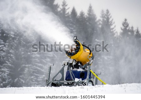 Snow cannon machine producing artificial snow on ski slope, preparation for winter season in mountain resort with forest background.