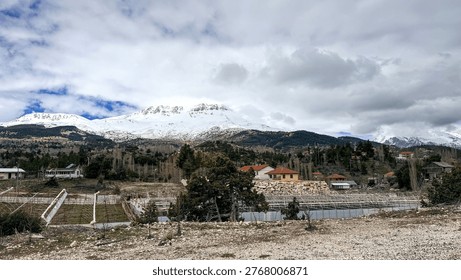 Snow capped mountain range under dramatic clouds, rural valley with small houses and fields in the foreground, calm scenic landscape with cool winter tones