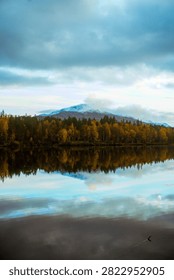 A snow capped mountain rises beyond a band of autumn trees along the Barduelva River, with low cloud and soft reflections spreading across the still water.