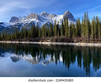 Snow capped mountains reflected in a mirror image on lake with blue sky and green spruce trees