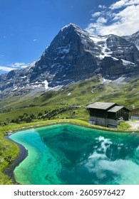 The snow capped north face of The Eiger with traditional Swiss chalet and turquoise pool from a mountain footpath. 
