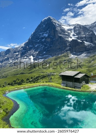 The snow capped north face of The Eiger with traditional Swiss chalet and turquoise pool from a mountain footpath. 