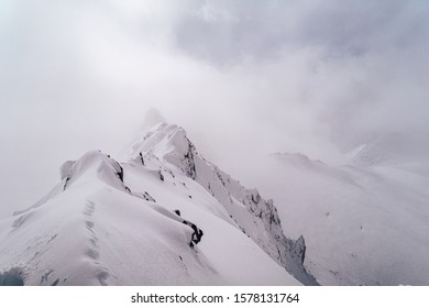 Snow Covered Alpine Ridge In Fog And Extreme Bad Weather. Alpine Mountian Landscape In Winter.