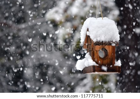 A snow covered bird house in winter with snowflakes falling down.