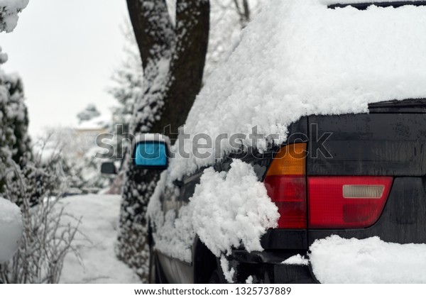 A snow covered\
black car photographed from the back - tail lights and blue rear\
view mirror visible; snow covered road, streetlamp, fence and trees\
visible to the side of the\
car.