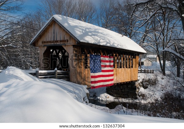 Snow covered bridge has an American flag\
draped on it to show patriotism. There are plenty of covered\
bridges in rural New Hampshire. These unique structures are\
favorite New England\
attractions.