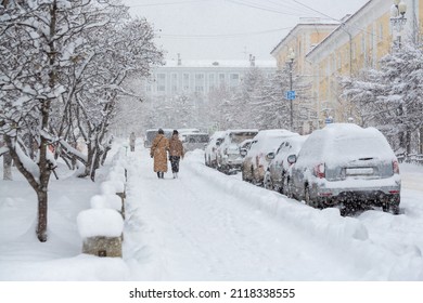 Snow covered city street during a heavy snowfall. Lots of snow on the sidewalk, cars and tree branches. Women walk around the winter city. Cold snowy weather. Magadan, Siberia, Russian Far East.