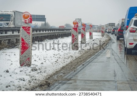 Snow covered highway at A7 E45 with traffic jam and red white construction barriers on a wet road during winter conditions under overcast sky.