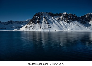 SNOW COVERED JAGGED PEAKSS WITH PARTIALLY EXPOSED ROCK WITH A BEAUTIFUL REFLECTION IN THE WATER AND A DEEP BLUE SKY NEAR SVALBARD NORWAY IN THE ARCTIC - Powered by Shutterstock