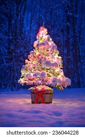 A Snow Covered Natural Spruce Christmas Tree With Illuminated Colorful Lights Sits In An Old Aged Wine Barrel Pot Outside In A Snowy Forest During The Winter Season At Night Time. 