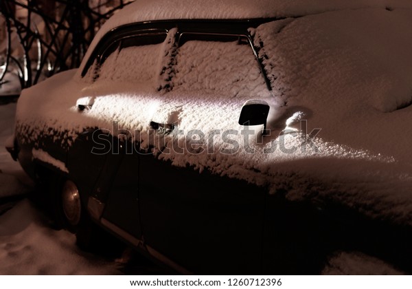 Snow covered old classic car illuminated by a ray of\
light at night. Old car covered with snow. Details of a car covered\
with snow