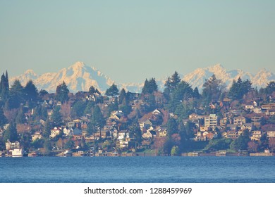 Snow covered peaks of the Olympic Mountains rise beautifully above and behind a Seattle hillside neighborhood on the shore of Lake Washington in view from Kirkland, WA on clear blue sky January day.