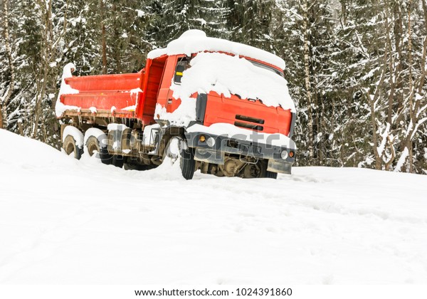 Snow covered the red truck left in the winter by\
the forest.