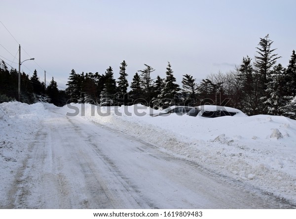 snow
covered road after snow plow has passed with tall snowbank on the 
and snow covered cars parked on the side of the
road