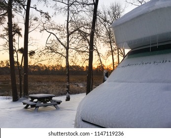 Snow Covered RV Camper At Sunrise Parked For Some Winter Camping At A Campground With A Picnic Table In The Background.