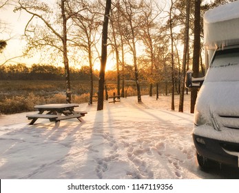 Snow Covered RV Camper At Sunrise Enjoying Some Winter Camping At A Campground With A Picnic Table In The Background.