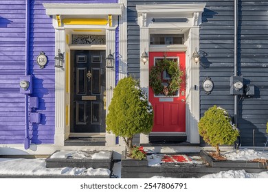 A snow covered sidewalk view of two doors to colorful wooden houses. Once has a bright red door with a Christmas wreath and the other is a solid black metal door with yellow trim and decorative trim. - Powered by Shutterstock