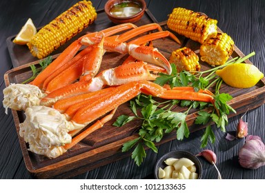 Snow Crab Legs Served With Melted Butter In Clay Bowls, Garlic Cloves, Lemon Slices, Grilled Corn In Cobs And Fresh Parsley On Wooden Cutting Boards, Horizontal View From Above, Close-up