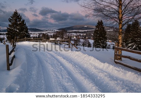 Snow drifts on road through twilight small and quiet winter alpine village, Voronenko, Carpathian, Ukraine. Night countryside hills, groves and farmlands in remote alpine village.