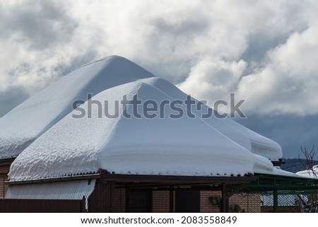 Snow drifts on the roof after the snowfall. In the background, the sky is covered with gray clouds. Roofs of the house, garage and shed in the snow. Three snow pyromids