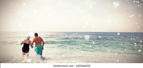 Snow Falling Against Rear View Of Senior Couple Holding Hands And Walking In Sea