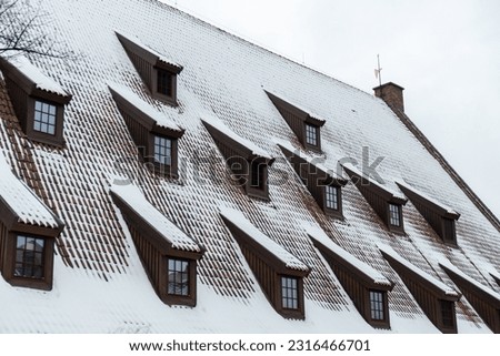 Snow falling on the roofs of the city. Snow-covered brown metal tiles roof of European house with windows. Rooftop covered