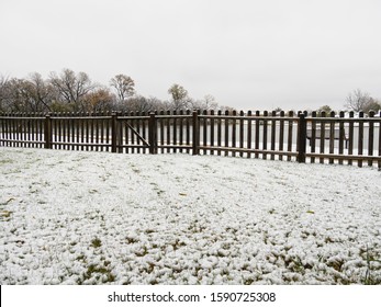 Snow Flurries Covering A Patch Of Grass In A Backyard, With A Wooden Fence In The Background