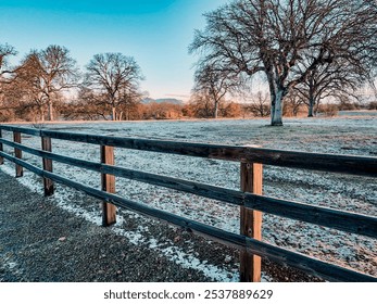 Snow and frost-covered field with bare trees, a split rail wooden fence in the foreground, and distant hills under a crisp blue sky, evoking a serene winter morning - Powered by Shutterstock