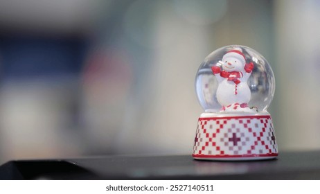 A snow globe featuring a cheerful snowman, set against a blurred background. The base has a red and white checkered pattern with green accents, evoking festive holiday spirit. - Powered by Shutterstock