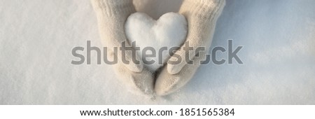Snow heart in hands. Human hands in warm beige gloves with snowy heart against snow background. I love winter or St.Valentine's Day romantic creative concept. Panorama banner with free space for text