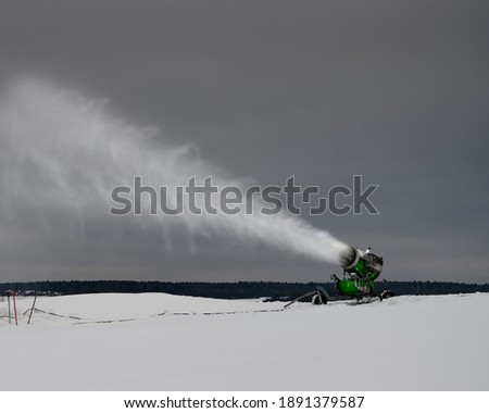 Snow making machine on the ski slope in winter. Spraying snow powder. White stream of snow on the background of the cloudy sky.
