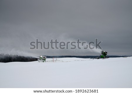 Snow making machine on the ski slope in winter. Spraying snow powder. White stream of snow on the background of the cloudy sky.