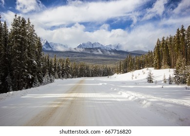 Snow On Small Narrow And Windy Mountain Road Through Forest With High Peaks Behind, Banff National Park, Canada